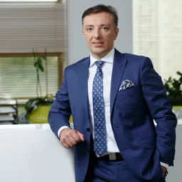 Portret elegancko ubranego mężczyzny w niebieskim garniturze, opierającego się o białe biurko w nowoczesnym biurze z roślinami w tle.