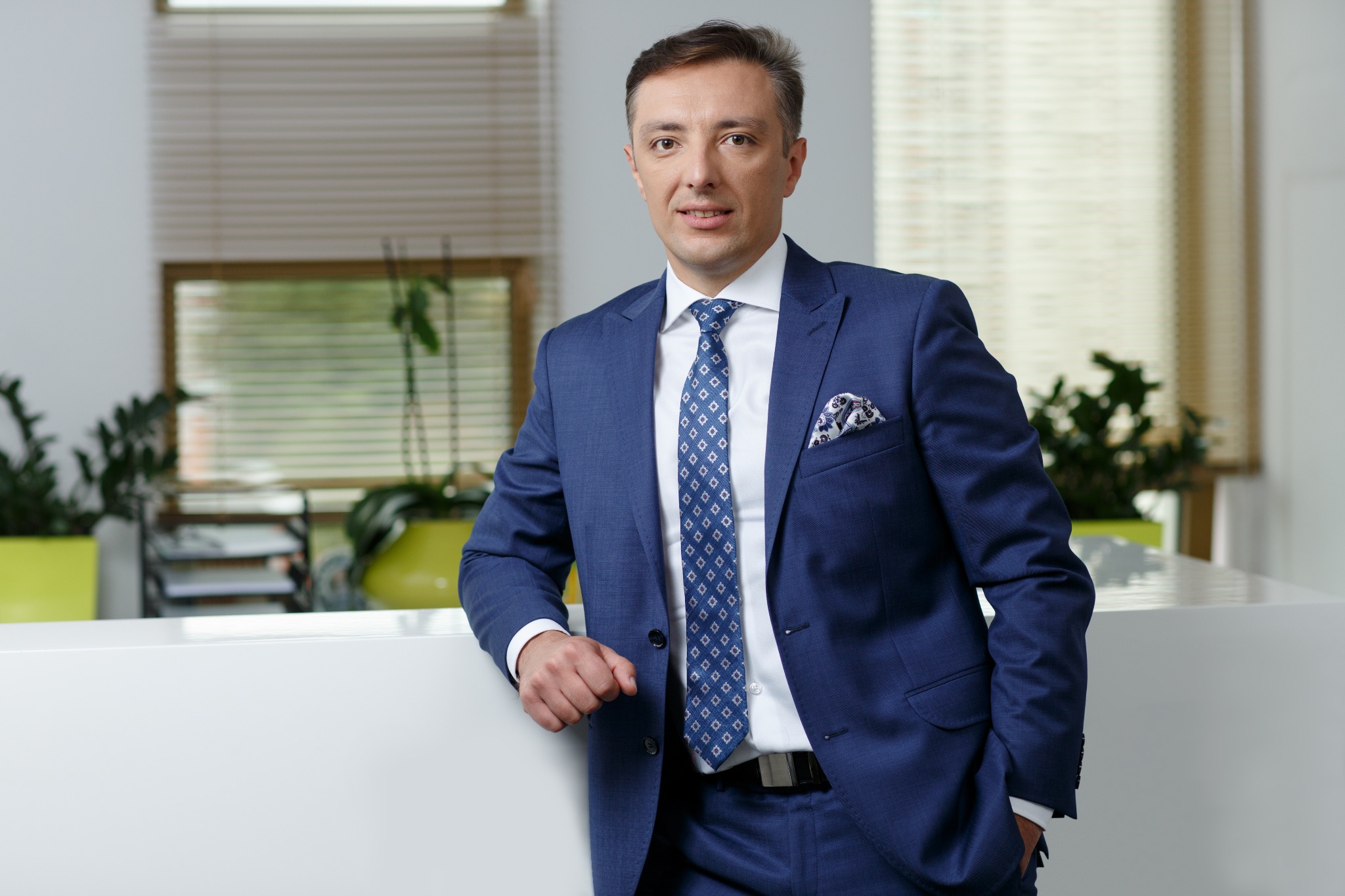 Portret elegancko ubranego mężczyzny w niebieskim garniturze, opierającego się o białe biurko w nowoczesnym biurze z roślinami w tle.