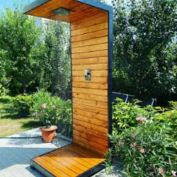 Nowoczesny prysznic zewnętrzny z drewnianymi panelami i czarną metalową ramą, otoczony zielenią i kwiatami w donicach, z widocznym strumieniem wody.