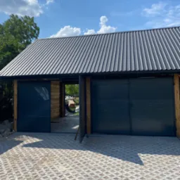 Nowoczesny garaż z dwiema przesuwnymi, ciemnymi bramami i metalowym dachem, wykończony drewnianymi belkami, z ażurową nawierzchnią z kostki betonowej na podjeździe, w tle zieleń drzew i błękitne...