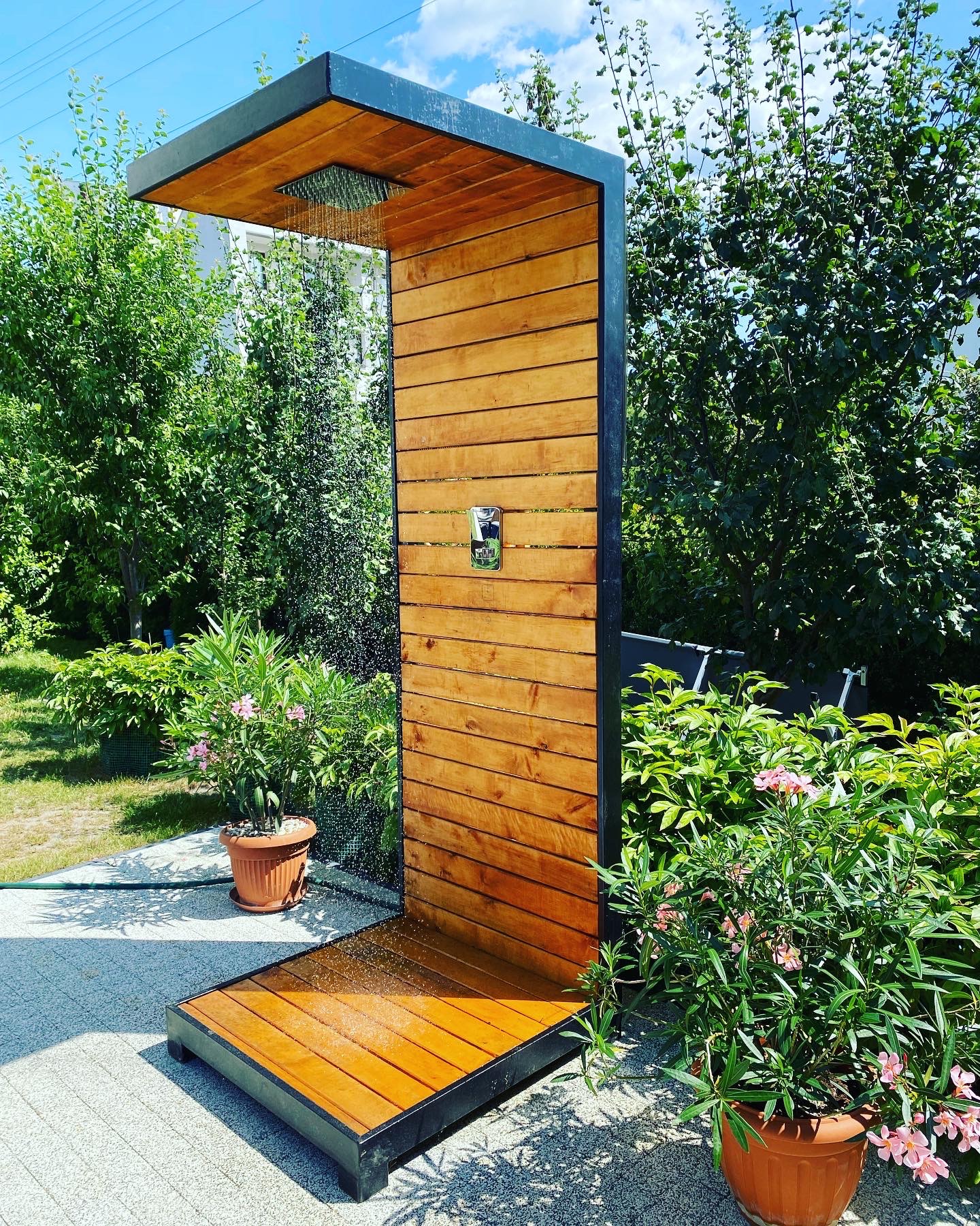 Nowoczesny prysznic zewnętrzny z drewnianymi panelami i czarną metalową ramą, otoczony zielenią i kwiatami w donicach, z widocznym strumieniem wody.