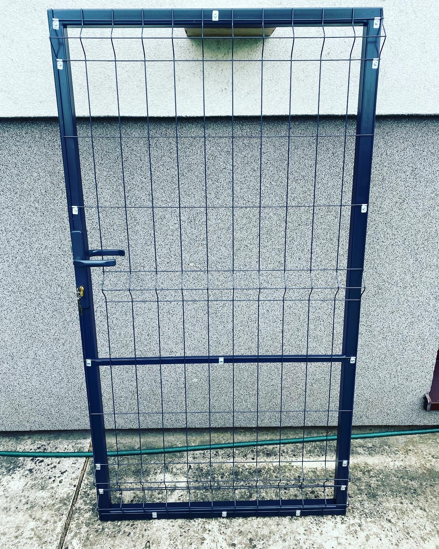 Czarna furtka ogrodzeniowa z wypełnieniem panelowym, z zamontowaną klamką i zamkiem, oparta o szarą, teksturowaną ścianę budynku, widoczny fragment zielonego węża ogrodowego na betonowej posadzce.