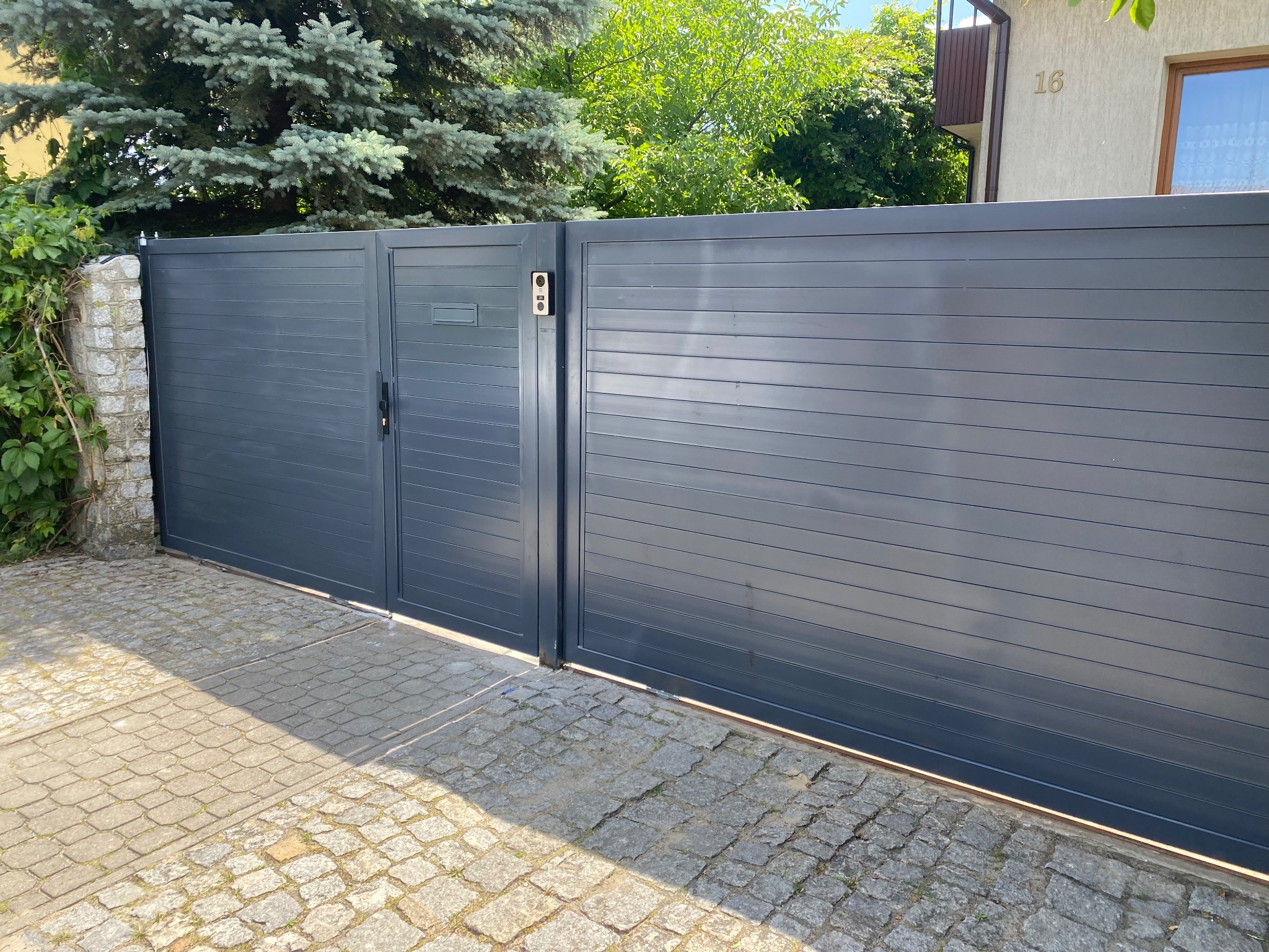 Nowoczesna, przesuwna brama wjazdowa w kolorze grafitowym z poziomymi panelami, zamontowana na posesji z brukowanym podjazdem i zielenią w tle.