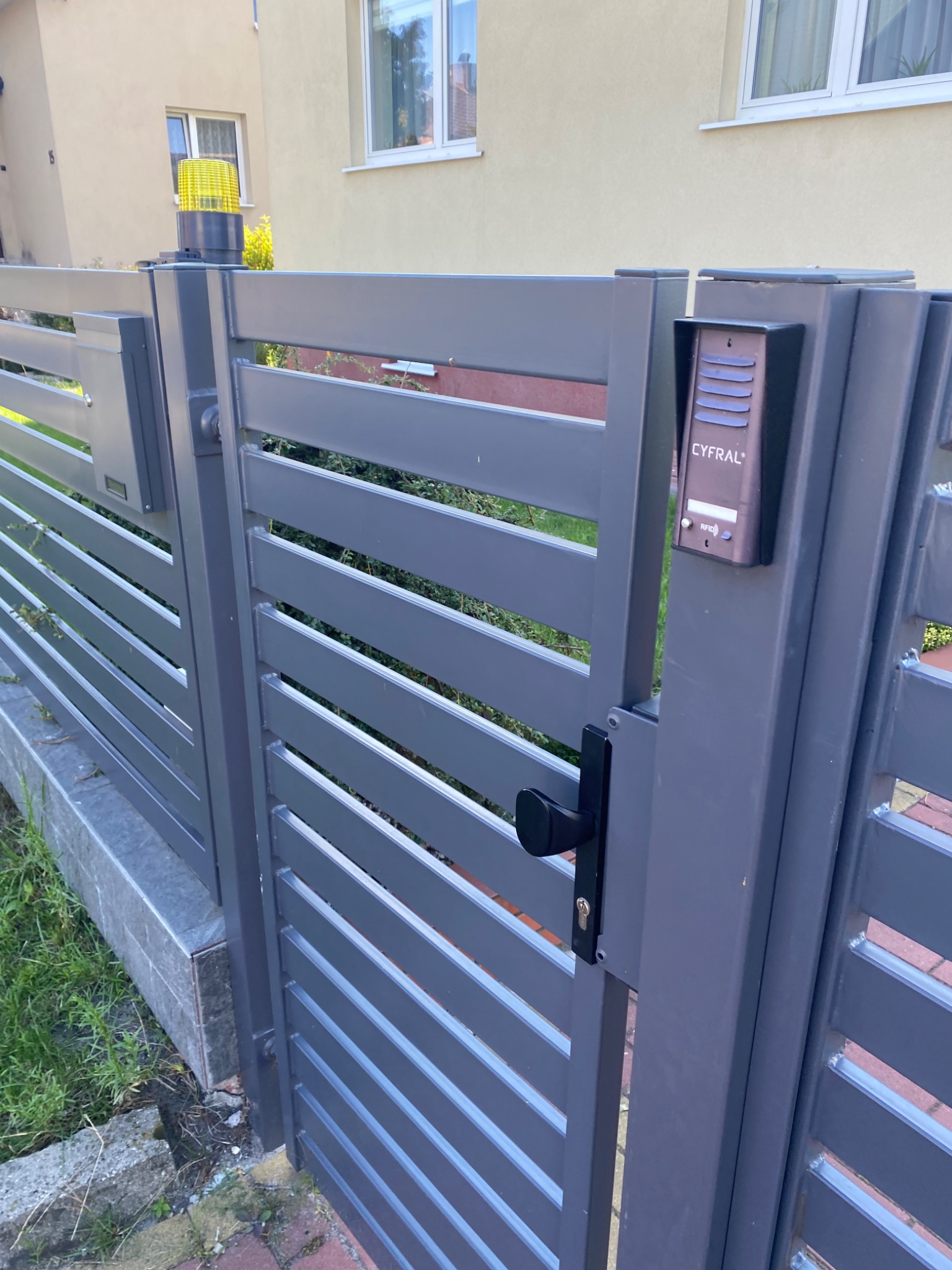 Nowoczesna brama wjazdowa z poziomych, szarych paneli, wyposażona w domofon Cyfral i skrzynkę na listy, z sygnalizatorem świetlnym na słupku, na tle jasnego budynku.