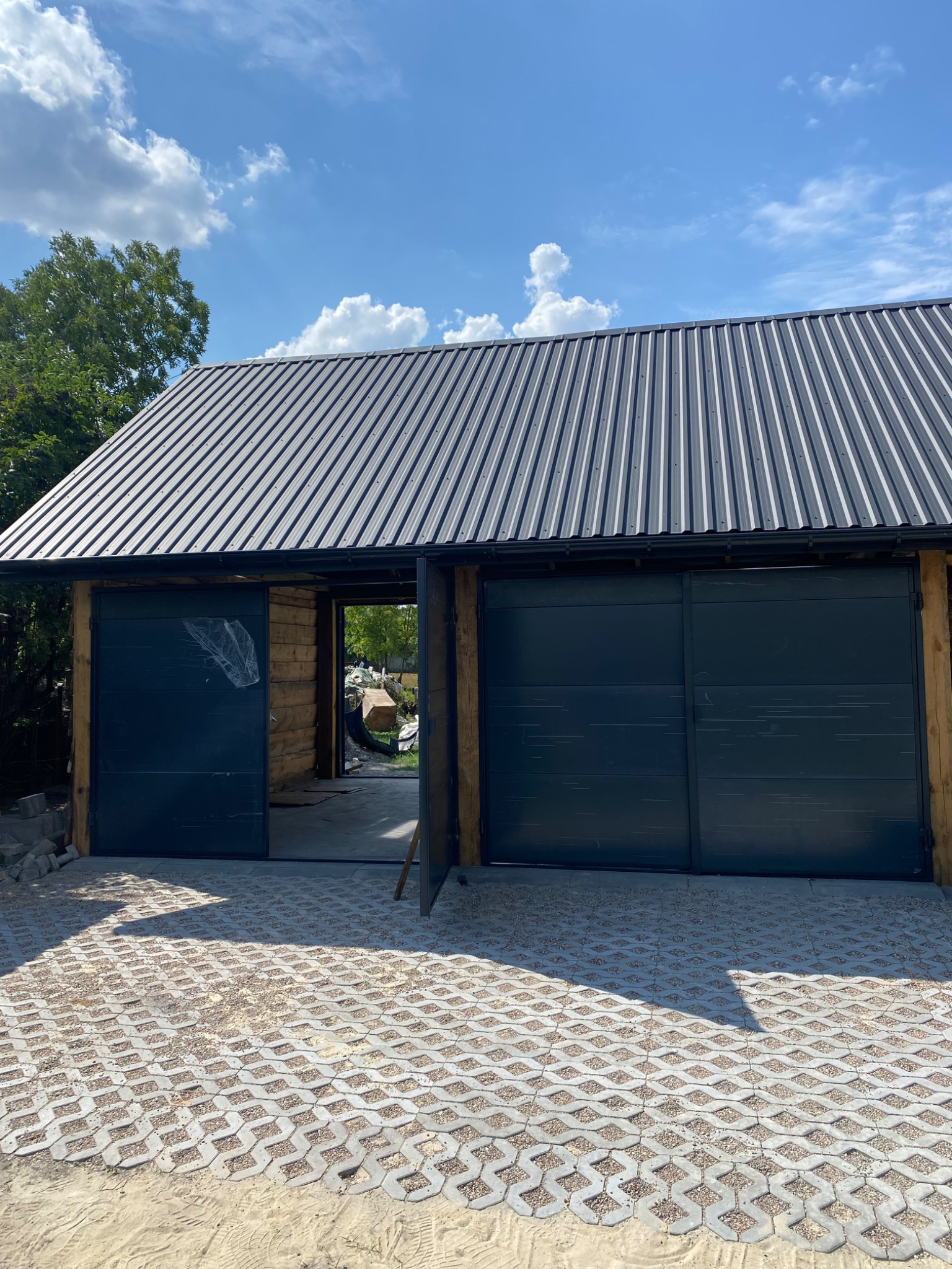 Nowoczesny garaż z dwiema przesuwnymi, ciemnymi bramami i metalowym dachem, wykończony drewnianymi belkami, z ażurową nawierzchnią z kostki betonowej na podjeździe, w tle zieleń drzew i błękitne...