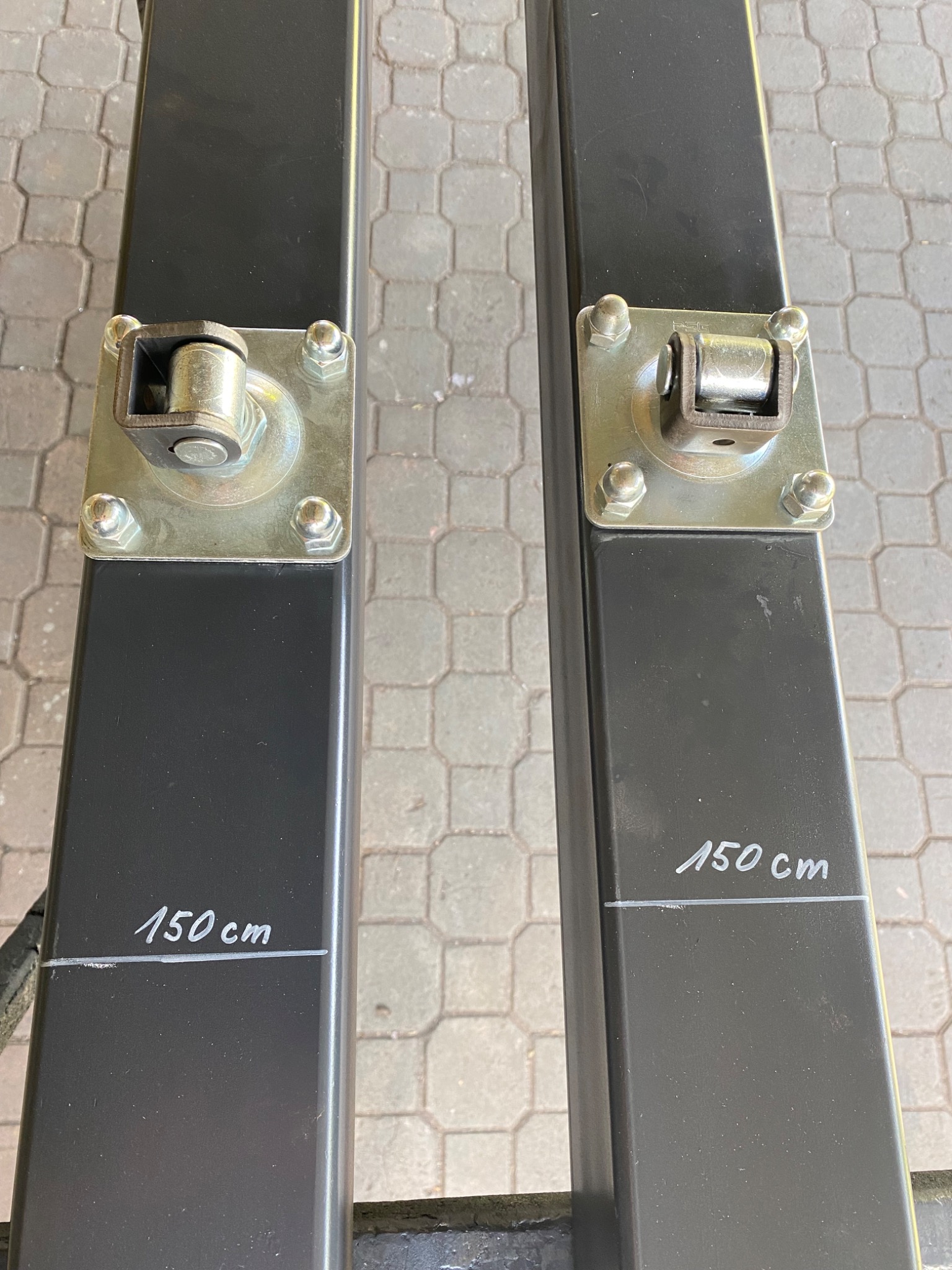 Dwa słupki z metalu pomalowane na czarno, z zaznaczoną wysokością 150 cm, zamocowane na metalowych podstawach z rolkami, widoczne na tle szarej kostki brukowej.