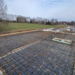 Zbrojenie stropu na czarnej folii z widocznymi elementami szalunku i rurami wentylacyjnymi, w tle dom w stanie surowym zamkniętym i zimowy krajobraz.