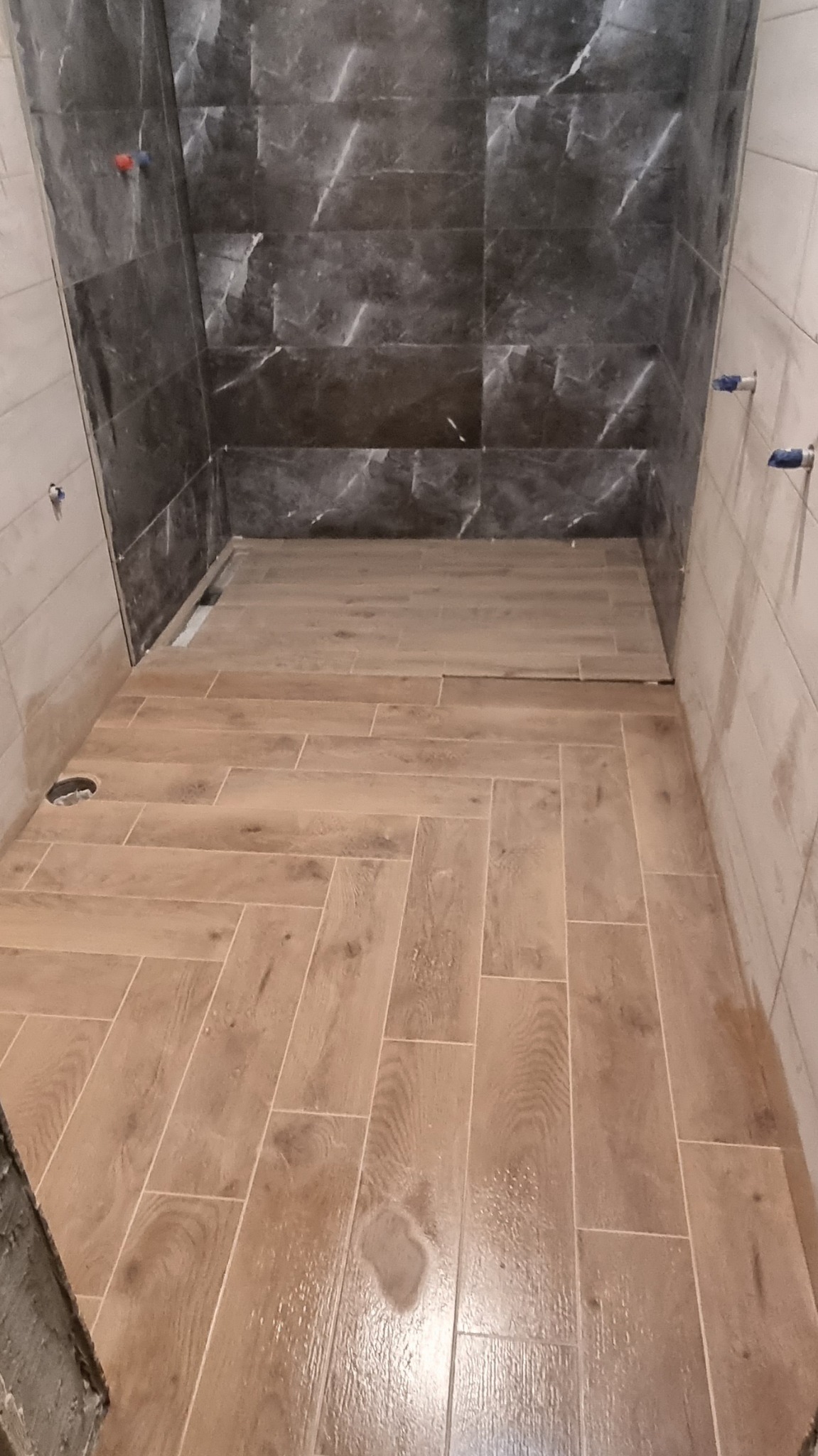 Ułożona podłoga z płytek imitujących drewno w jodełkę w łazience z częściowo wykończonymi ścianami, widoczne przyłącza wodne i odpływ w podłodze.