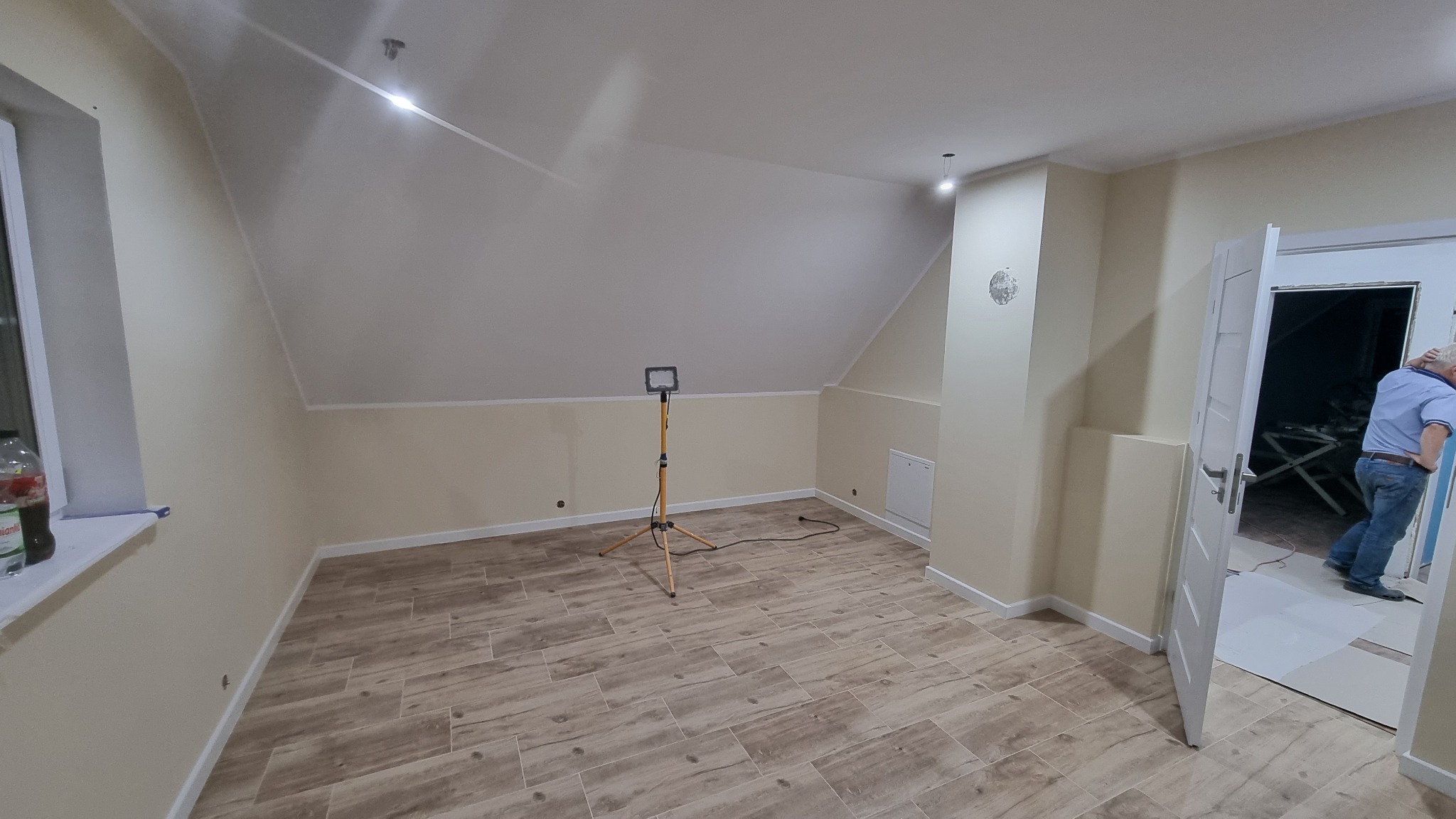 Poddasze po remoncie z nową podłogą imitującą drewno, świeżo pomalowanymi ścianami w odcieniach beżu i bieli, oświetlone lampą na statywie, z widocznym wejściem i fragmentem sylwetki osoby wewnątrz.