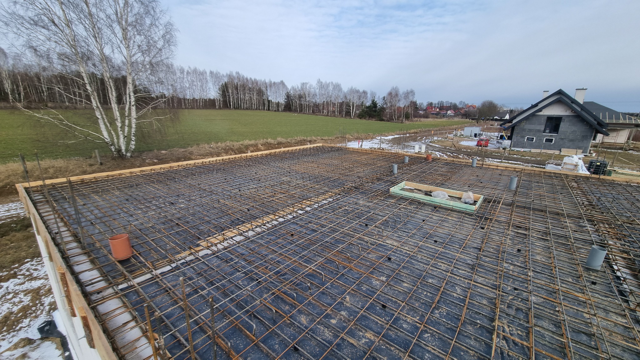 Zbrojenie stropu na czarnej folii z widocznymi elementami szalunku i rurami wentylacyjnymi, w tle dom w stanie surowym zamkniętym i zimowy krajobraz.