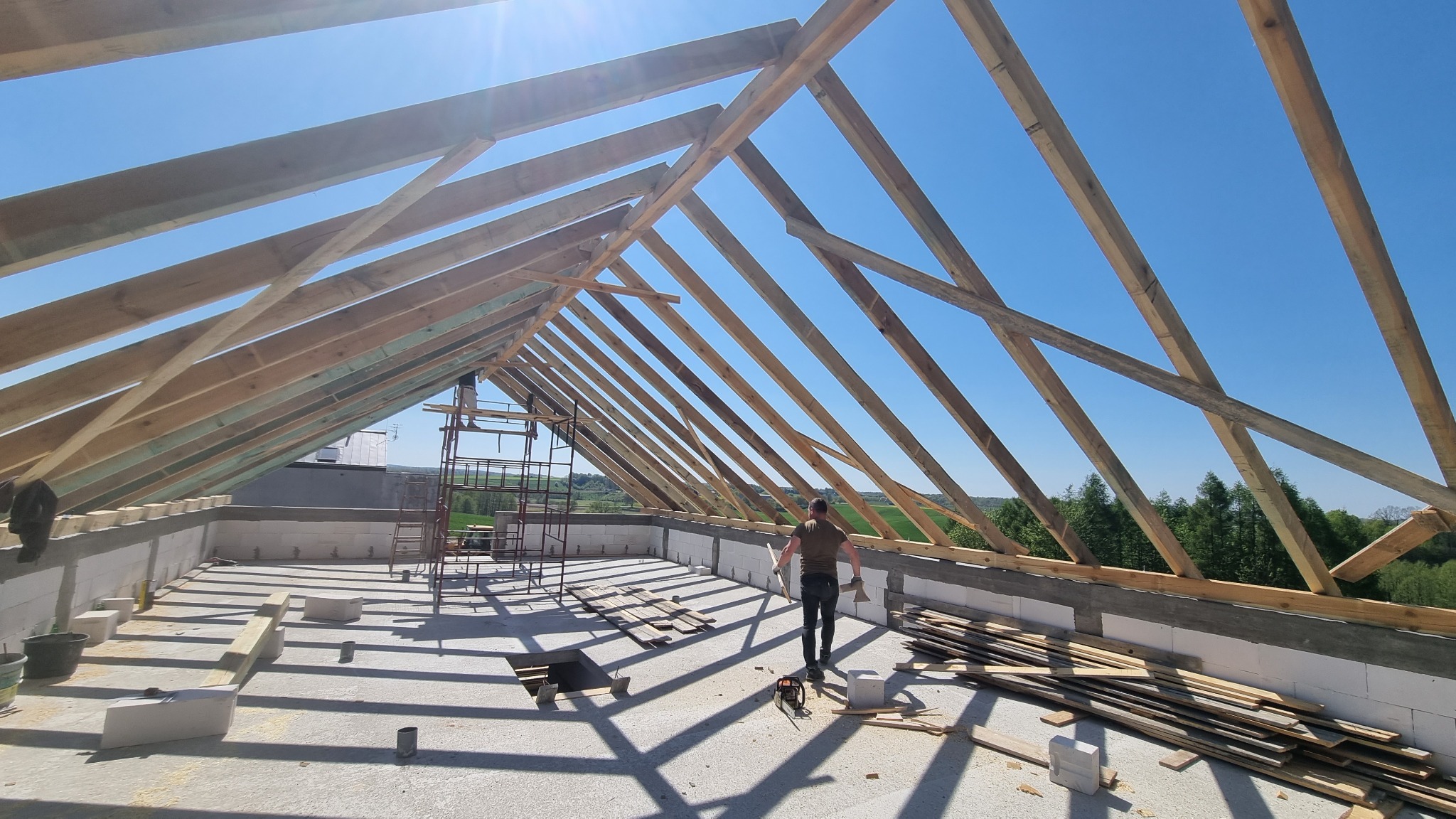 Konstrukcja drewniana dachu w trakcie budowy, widoczny szkielet z belek na tle błękitnego nieba, mężczyzna na tle krajobrazu, elementy rusztowania.