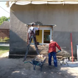 Pracownicy podczas wymiany okna w budynku z szarą, chropowatą elewacją. Jeden pracownik na drabinie uszczelnia okolice okna, drugi zbiera gruz do taczki. Widoczne drewniane elementy konstrukcji dachu.