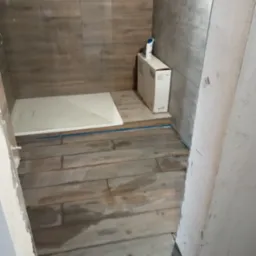 Montaż kabiny prysznicowej: widoczny brodzik, częściowo ułożona podłoga z płytek imitujących drewno, karton po ceramice i niebieska taśma malarska.