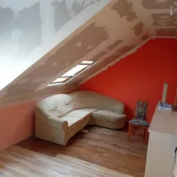 Poddasze w trakcie prac wykończeniowych: ściany z nałożoną gładzią, fragment pomalowanej na pomarańczowo ściany, ułożone panele podłogowe, narożna sofa i krzesło w tle.