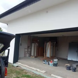 Świeżo pomalowana elewacja budynku z otwartym garażem, widoczne puszki z farbą, samochód z otwartym bagażnikiem i elementy instalacji CO wewnątrz garażu.