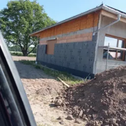 Dom w trakcie ocieplania elewacji: widoczne szare płyty izolacyjne z otworami, częściowo zamontowane, ceglana ściana powyżej, rusztowanie przy oknie, pryzma ziemi na pierwszym planie, drzewo w tle.