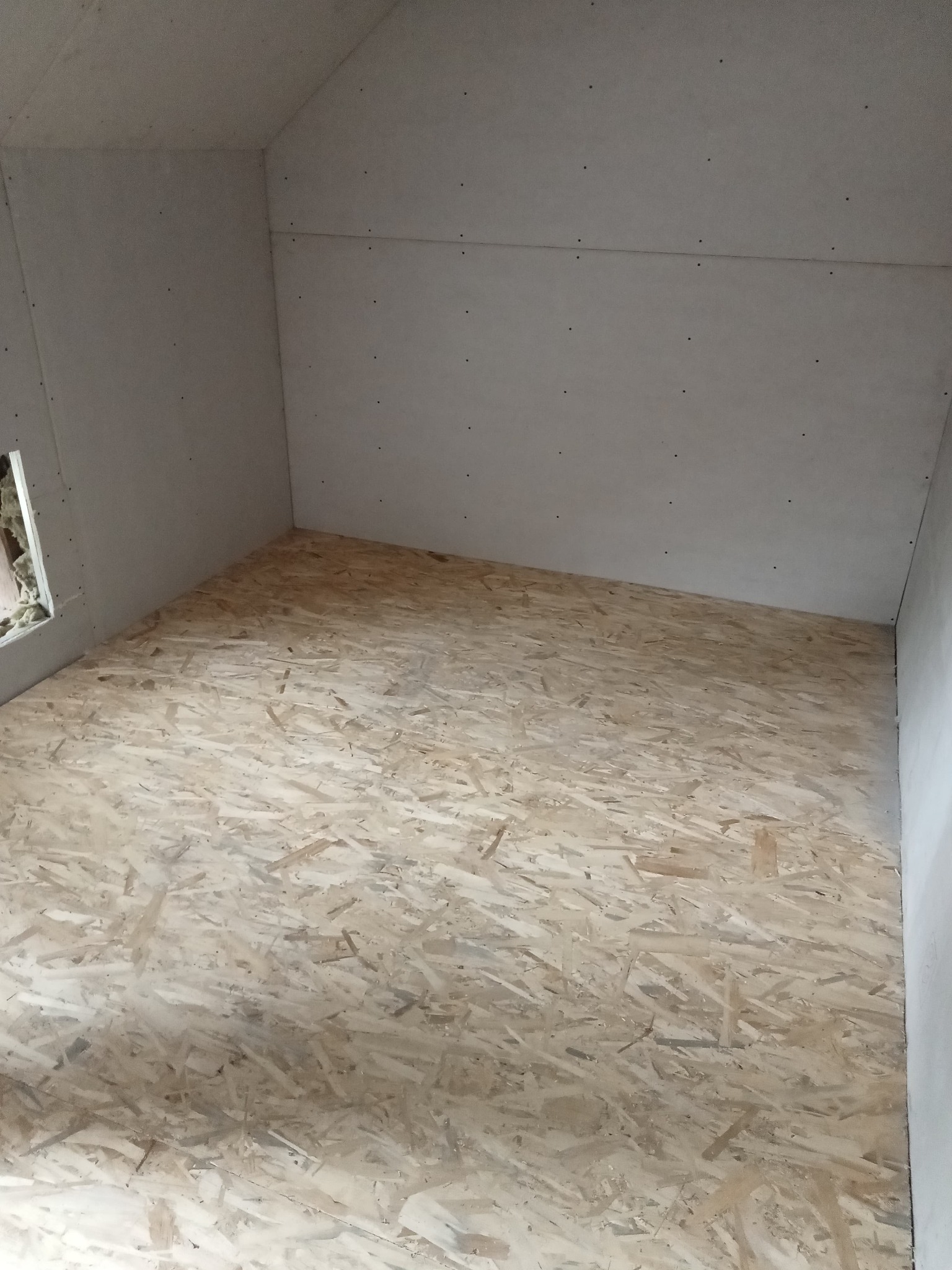 Pokój na poddaszu w trakcie wykańczania: ściany obłożone płytami kartonowo-gipsowymi, podłoga wyłożona płytami OSB, widoczny otwór okienny z izolacją.