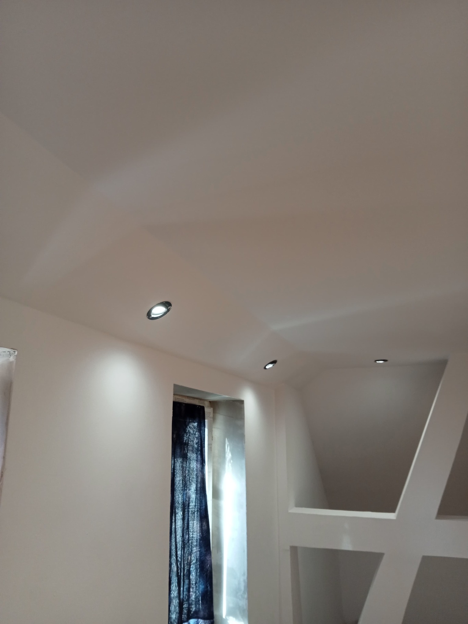 Gładki, biały sufit podwieszany z zamontowanymi punktowymi lampami LED, widoczne fragmenty ścian i okna z zasłoną, element dekoracyjny na ścianie.