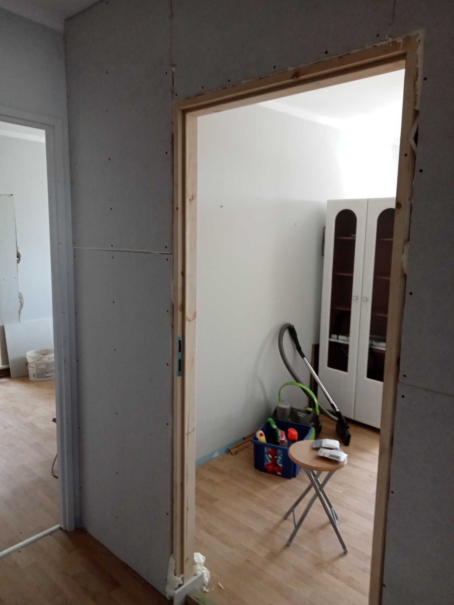 Montaż ościeżnicy drewnianej w ścianie z płyt kartonowo-gipsowych, widoczny etap prac wykończeniowych, narzędzia i akcesoria budowlane w tle.