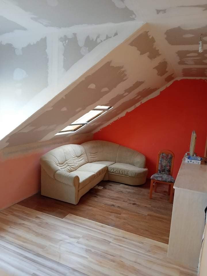 Poddasze w trakcie prac wykończeniowych: ściany z nałożoną gładzią, fragment pomalowanej na pomarańczowo ściany, ułożone panele podłogowe, narożna sofa i krzesło w tle.