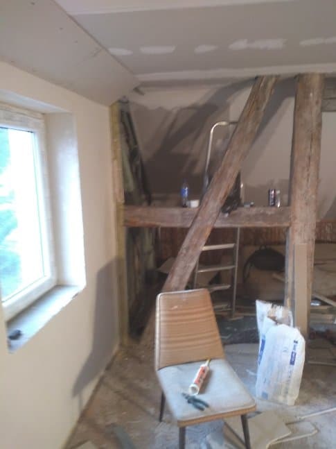 Wnętrze remontowanego poddasza z widoczną konstrukcją drewnianą, częściowo zamontowaną płytą karton-gips na suficie, oknem po lewej stronie i krzesłem z tubą silikonu i szczypcami na siedzisku.