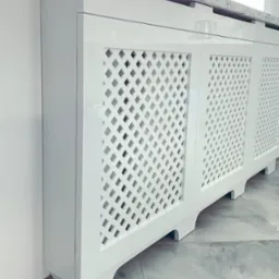 Biała, lakierowana obudowa grzejnika z ażurowym frontem w kształcie rombów i marmurowym blatem, dopasowana do wnętrza.