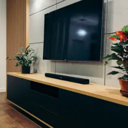 Nowoczesny salon z czarną szafką RTV z drewnianym blatem, na której stoi głośnik soundbar i rośliny doniczkowe, a nad nią wisi telewizor na ścianie z betonowych płyt. Dekoracyjna ściana...