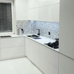 Nowoczesna, minimalistyczna kuchnia w bieli z blatem i backsplashiem w czarno-białe plamki, czarna bateria i płyta grzewcza, metalowy czajnik przy oknie z żaluzjami.