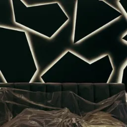 Ciemne wnętrze z podświetloną, geometryczną dekoracją ścienną nad wezgłowiem łóżka przykrytego folią ochronną.