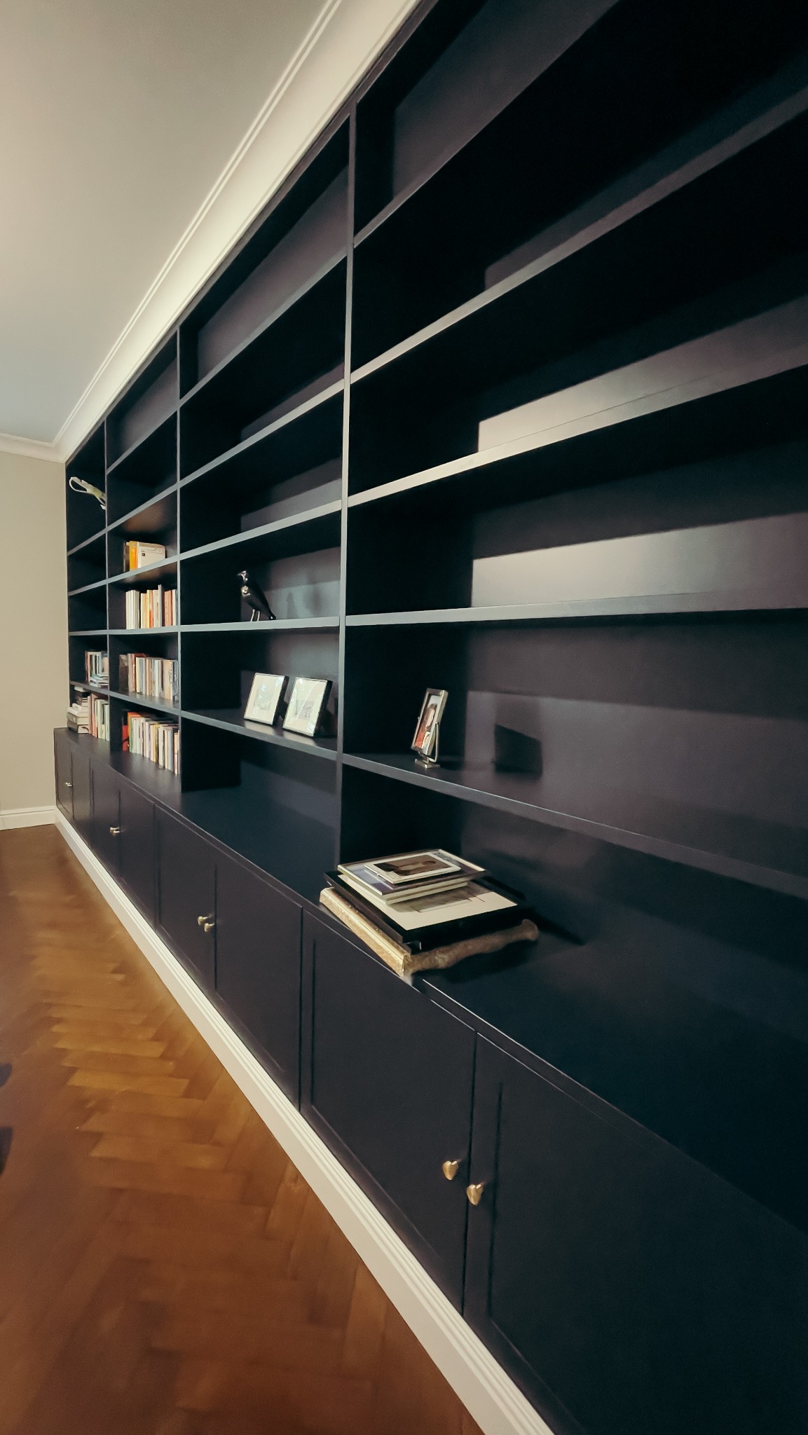 Długa, zabudowana, ciemna biblioteczka z szafkami w dolnej części, z książkami i dekoracjami na półkach, na podłodze parkiet ułożony w jodełkę.