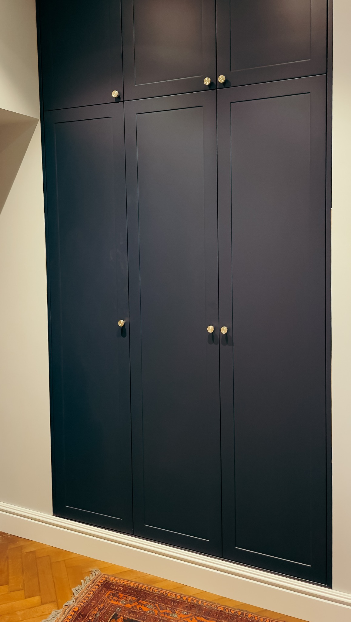 Ciemnogranatowa szafa wnękowa z trzema parami złotych gałek, widoczna część parkietu i dywan z orientalnym wzorem.