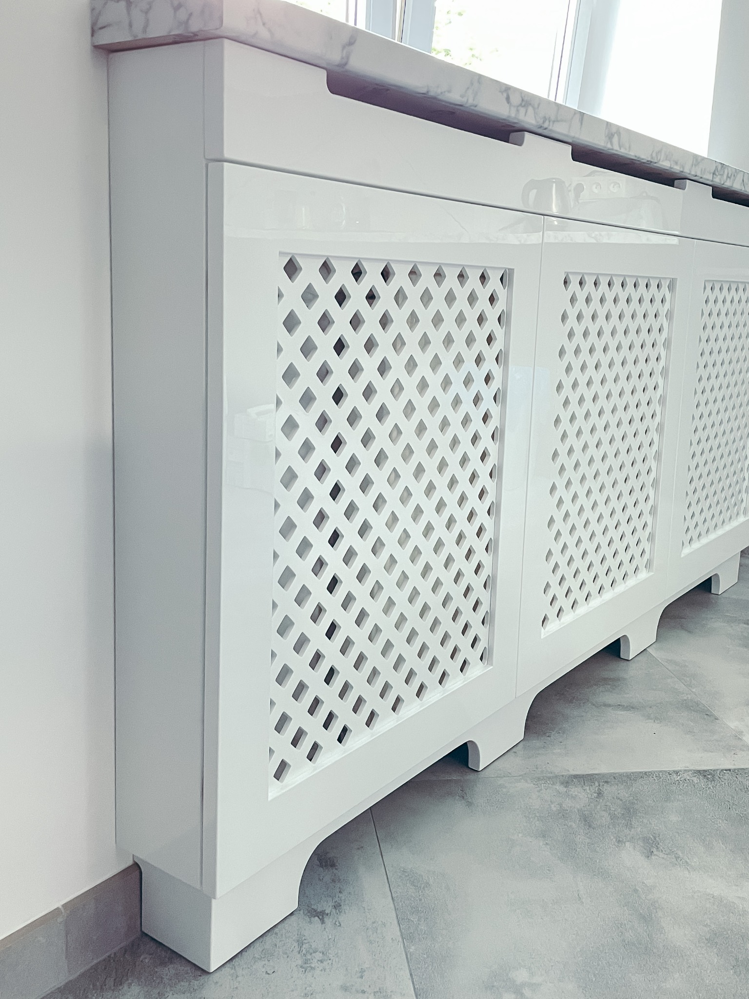Biała, lakierowana obudowa grzejnika z ażurowym frontem w kształcie rombów i marmurowym blatem, dopasowana do wnętrza.