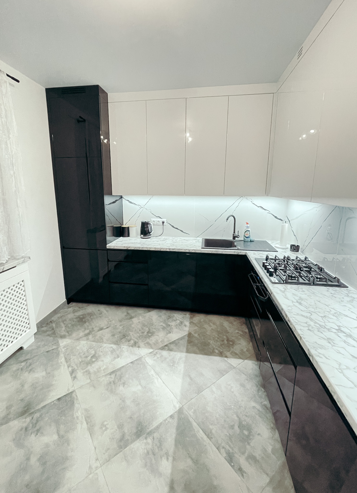 Nowoczesna kuchnia z białymi, błyszczącymi szafkami wiszącymi i czarnymi szafkami dolnymi, blatem imitującym marmur i szarą podłogą w geometryczny wzór.