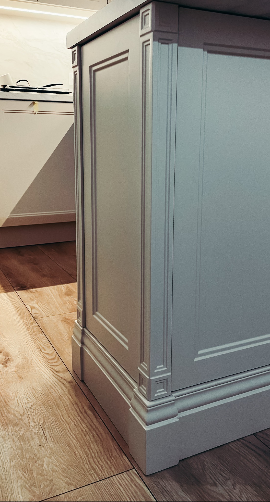 Detale wykończenia wyspy kuchennej w stylu klasycznym: frezowane panele, dekoracyjne listwy i drewniana podłoga w ciepłym odcieniu.