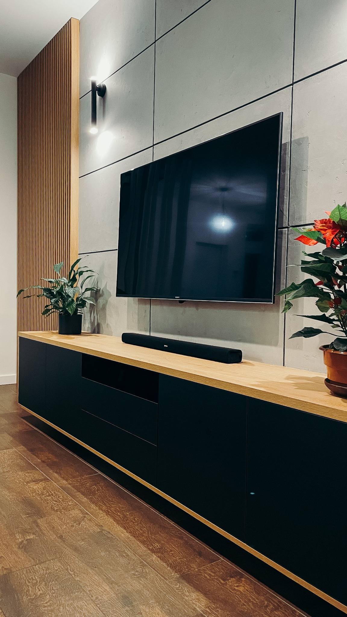 Nowoczesny salon z czarną szafką RTV z drewnianym blatem, na której stoi głośnik soundbar i rośliny doniczkowe, a nad nią wisi telewizor na ścianie z betonowych płyt. Dekoracyjna ściana...