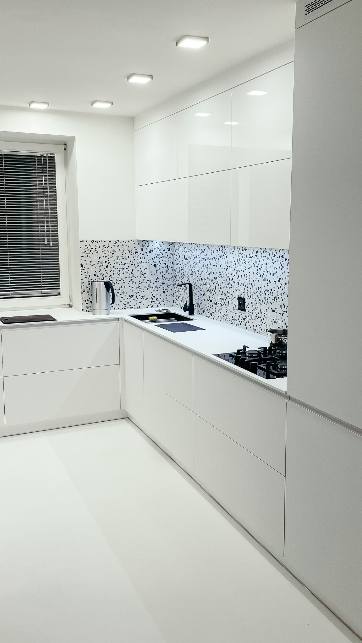 Nowoczesna, minimalistyczna kuchnia w bieli z blatem i backsplashiem w czarno-białe plamki, czarna bateria i płyta grzewcza, metalowy czajnik przy oknie z żaluzjami.