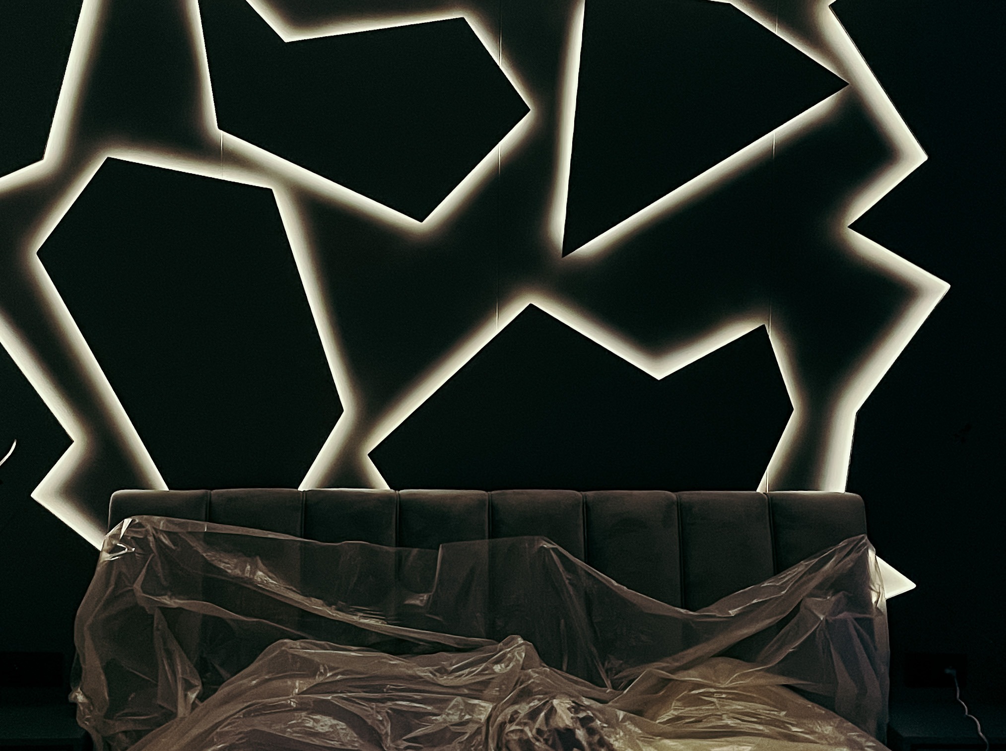 Ciemne wnętrze z podświetloną, geometryczną dekoracją ścienną nad wezgłowiem łóżka przykrytego folią ochronną.