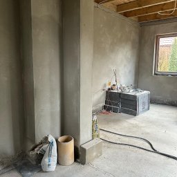 Bud-Pat - Surowe wnętrze w trakcie prac wykończeniowych: świeżo położone tynki na ścianach, widoczne worki z zaprawą, narzędzia i kable na podłodze, drewniany sufit i okno z widokiem na zieleń.