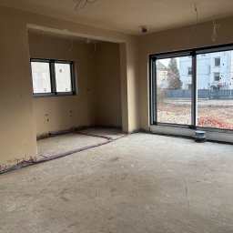 Bud-Pat - Surowe wnętrze nowo wybudowanego domu przed wykończeniem: ściany pokryte wstępnym tynkiem, widoczne instalacje elektryczne i rury grzewcze wzdłuż podłogi, duże okno z widokiem na budynek mieszkalny.
