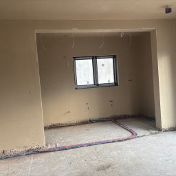 Bud-Pat - Wnętrze w trakcie prac wykończeniowych: gładkie, beżowe ściany po nałożeniu gładzi, widoczne przewody elektryczne przy podłodze, okno w czarnej ramie i otwór drzwiowy bez drzwi.