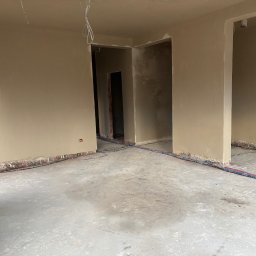 Bud-Pat - Wnętrze w trakcie prac wykończeniowych: świeżo otynkowane ściany w beżowym odcieniu, widoczne wystające przewody elektryczne i instalacje wodne na podłodze, surowy betonowy podkład.