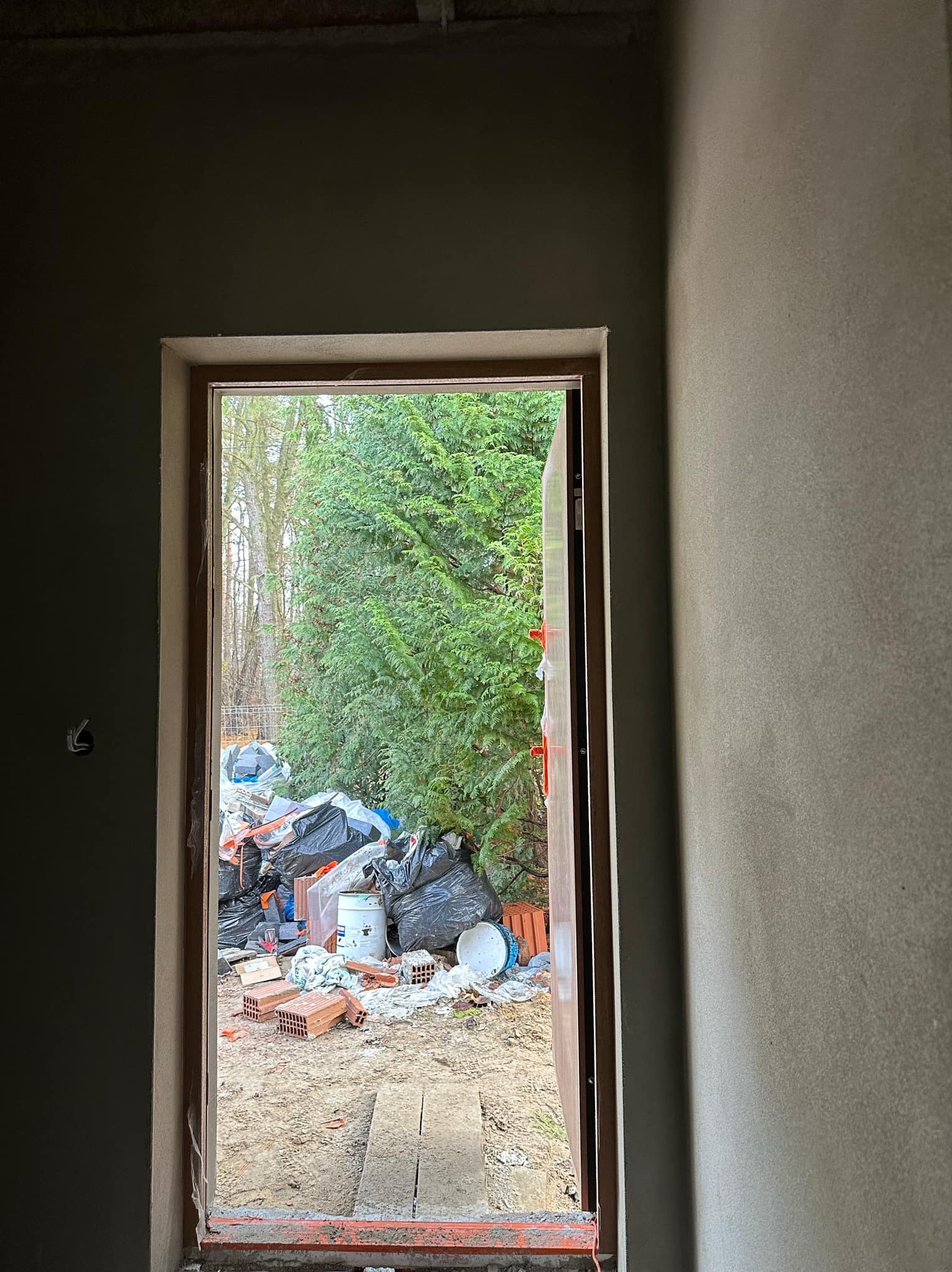Surowe wnętrze z widokiem na zewnątrz przez otwór drzwiowy, ściany pokryte świeżym tynkiem, na zewnątrz widoczne gruz i materiały budowlane oraz zieleń.