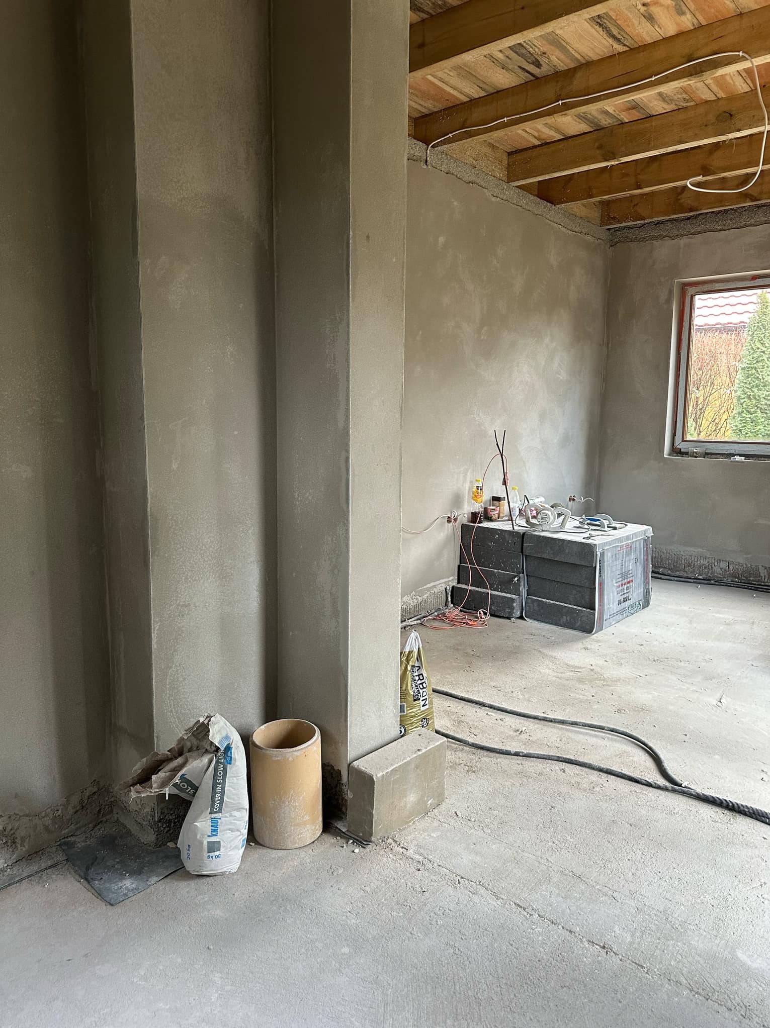 Surowe wnętrze w trakcie prac wykończeniowych: świeżo położone tynki na ścianach, widoczne worki z zaprawą, narzędzia i kable na podłodze, drewniany sufit i okno z widokiem na zieleń.