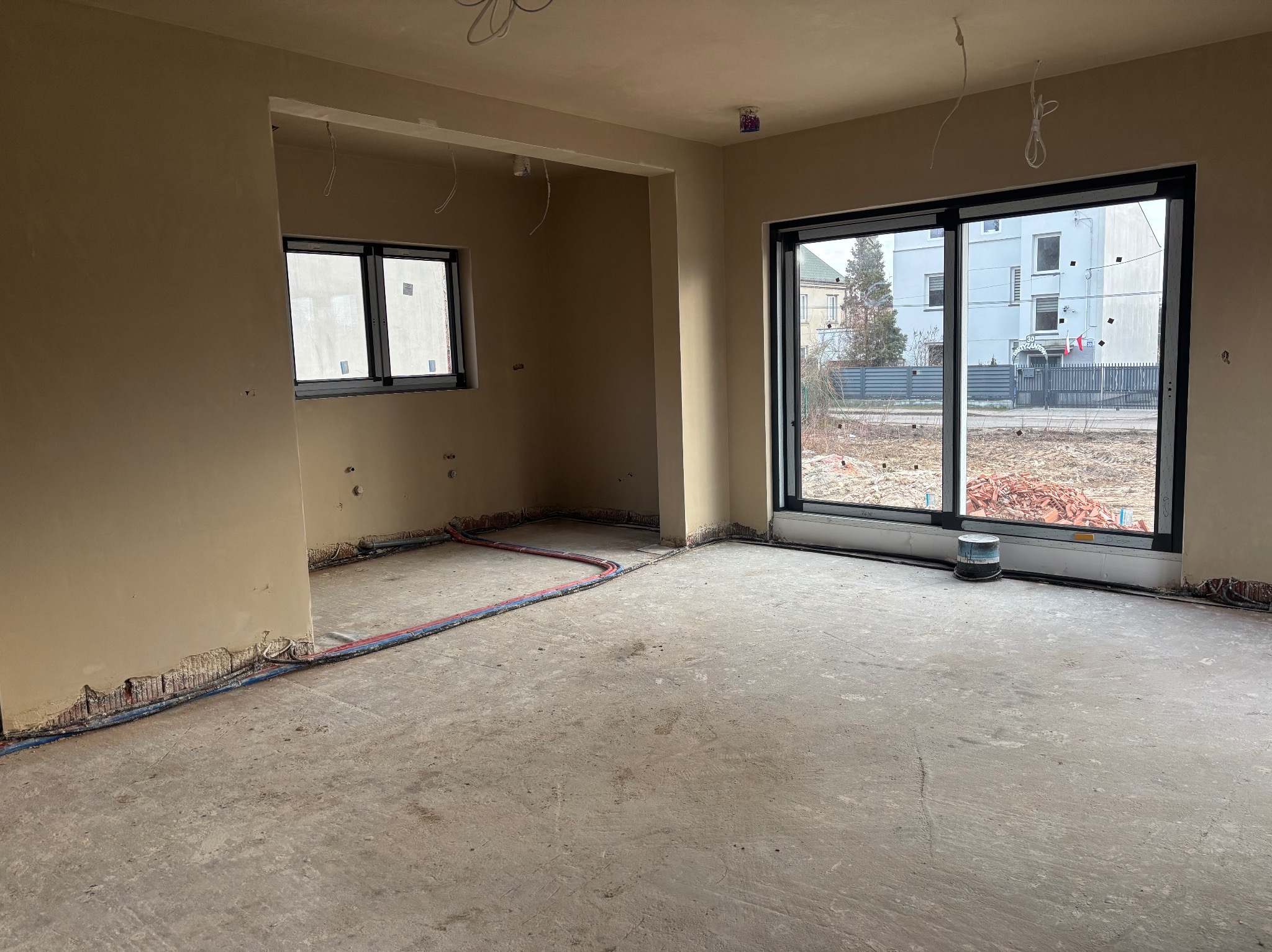 Surowe wnętrze nowo wybudowanego domu przed wykończeniem: ściany pokryte wstępnym tynkiem, widoczne instalacje elektryczne i rury grzewcze wzdłuż podłogi, duże okno z widokiem na budynek mieszkalny.