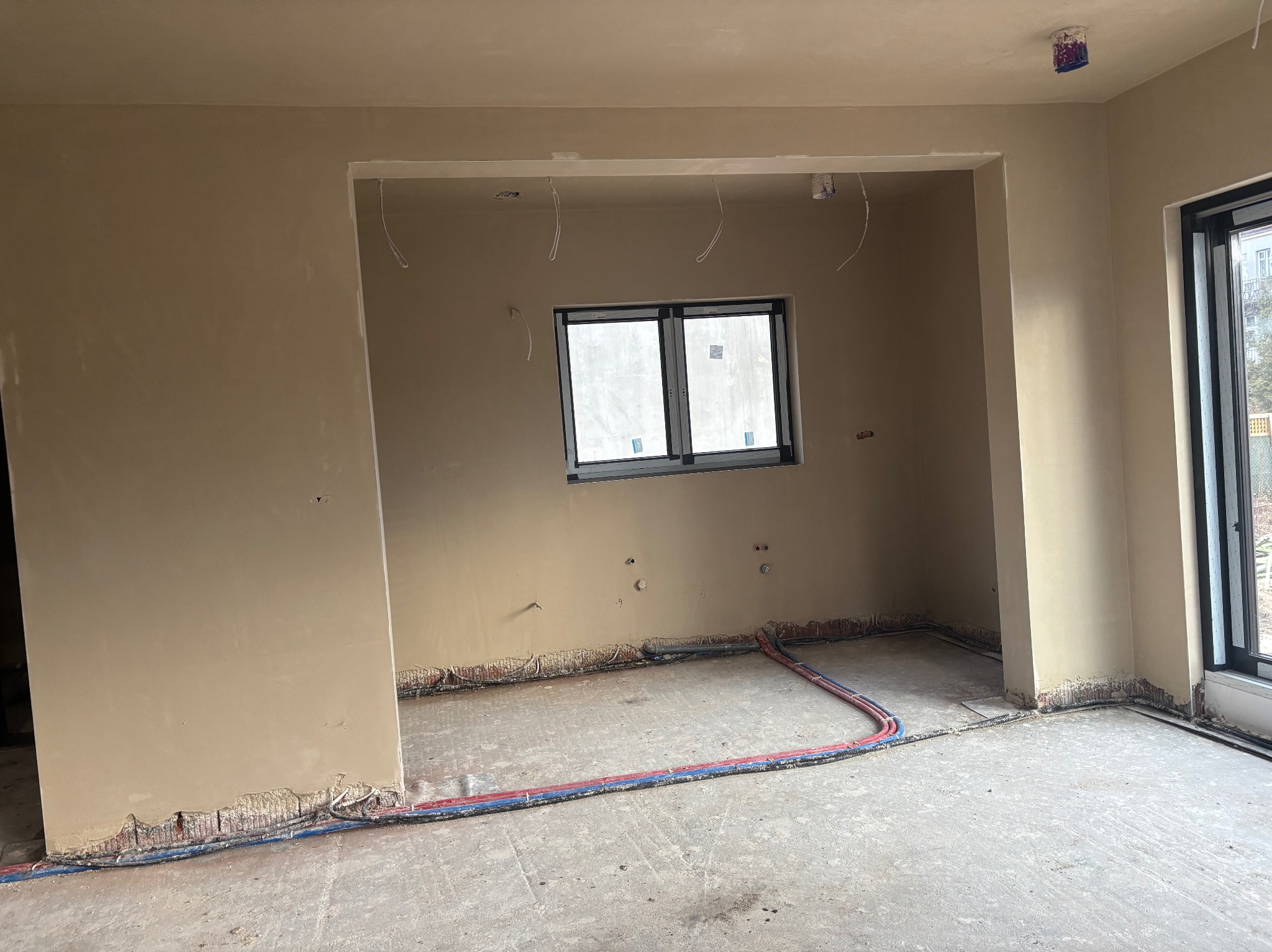Wnętrze w trakcie prac wykończeniowych: gładkie, beżowe ściany po nałożeniu gładzi, widoczne przewody elektryczne przy podłodze, okno w czarnej ramie i otwór drzwiowy bez drzwi.