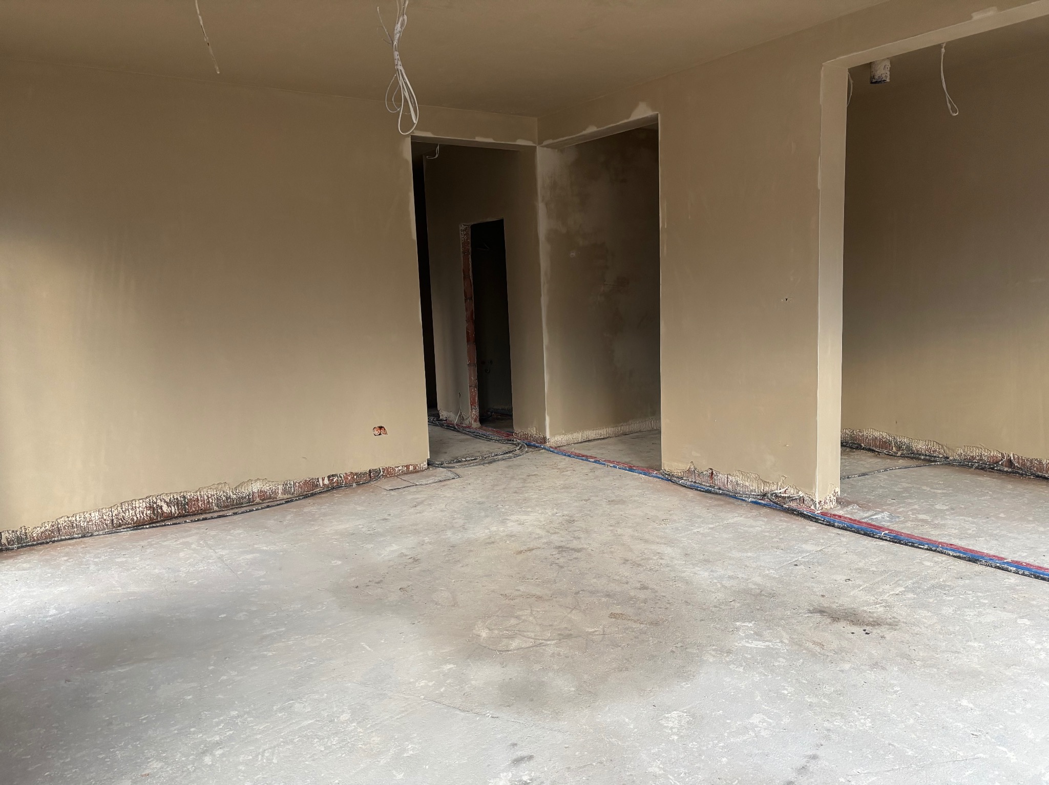 Wnętrze w trakcie prac wykończeniowych: świeżo otynkowane ściany w beżowym odcieniu, widoczne wystające przewody elektryczne i instalacje wodne na podłodze, surowy betonowy podkład.