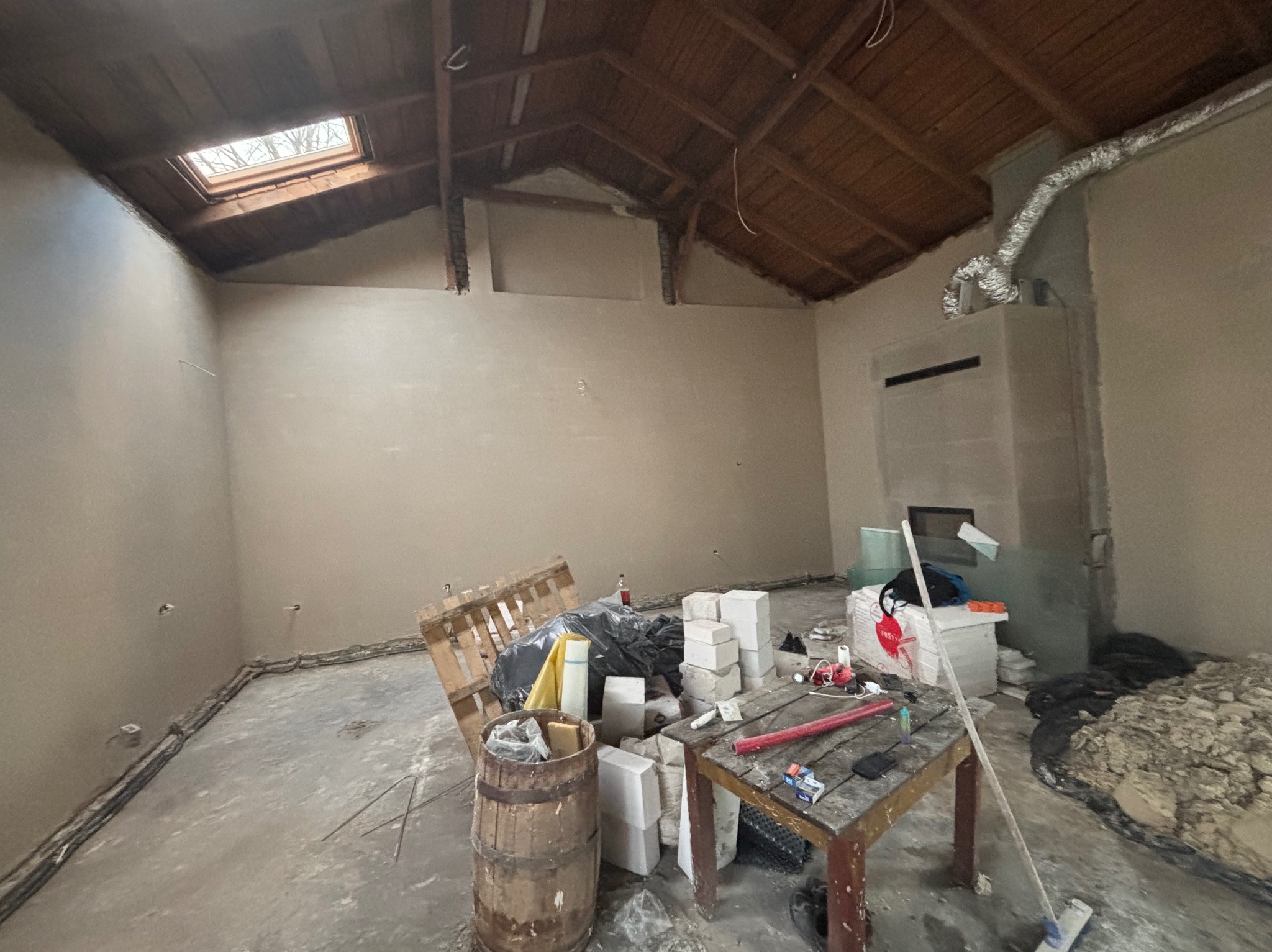 Wnętrze remontowanego poddasza z oknem dachowym, ściany pokryte świeżym tynkiem, widoczne materiały budowlane i narzędzia na podłodze oraz drewniana konstrukcja dachu.