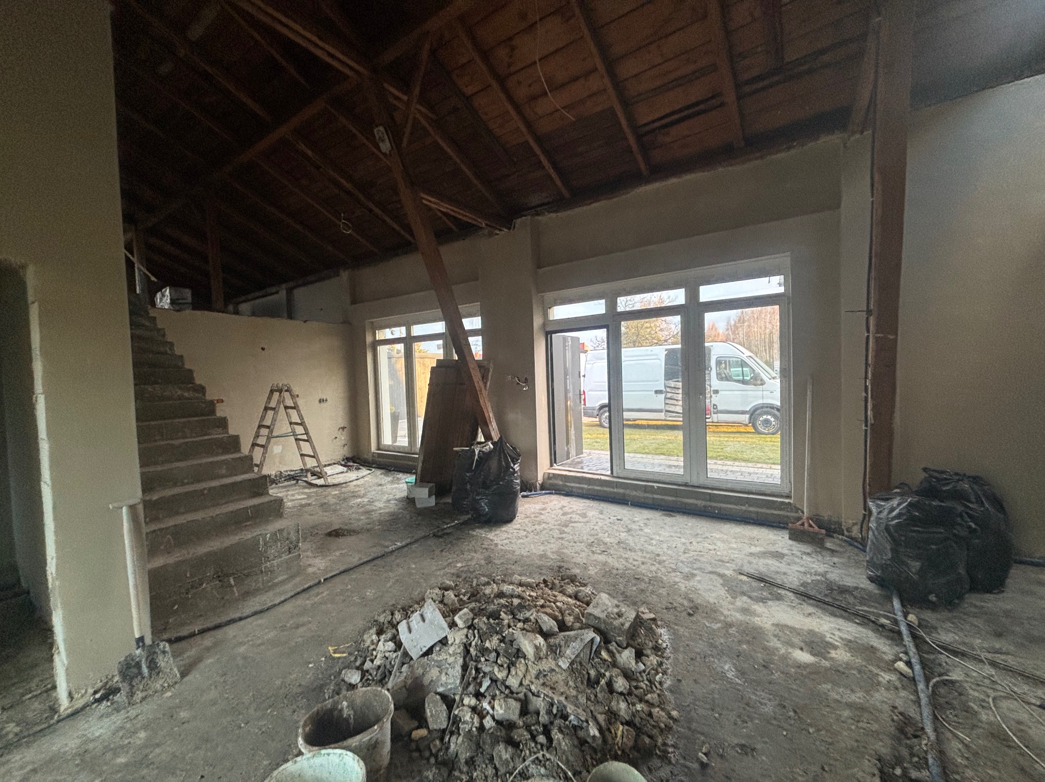 Wnętrze w trakcie remontu: surowe ściany, drewniane krokwie, prowizoryczne schody, gruz na podłodze, drabina i widok na zaparkowany biały van za oknem.