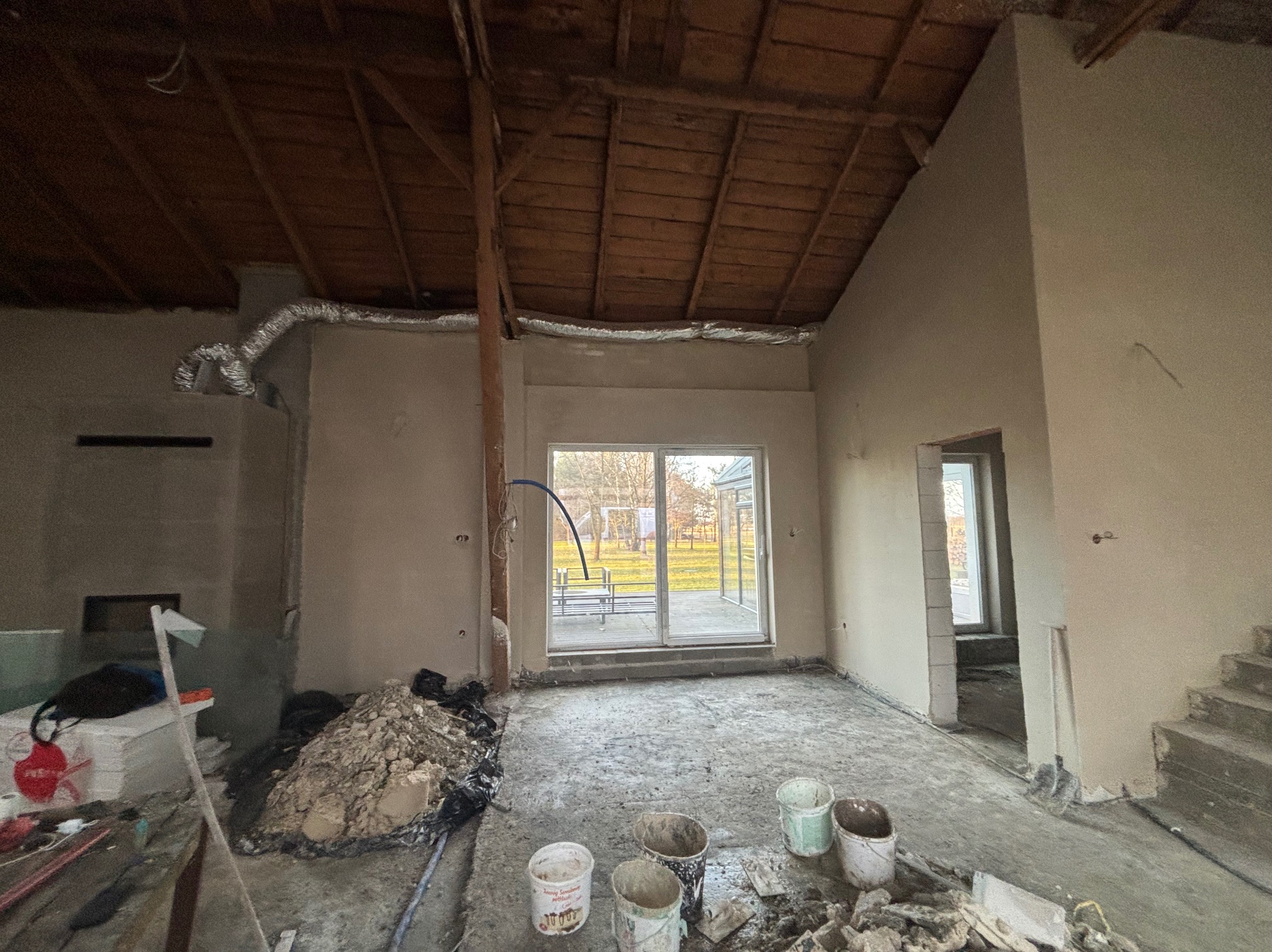 Wnętrze w trakcie prac wykończeniowych: świeżo otynkowane ściany, drewniany strop, widoczne wiadra i resztki materiałów budowlanych na podłodze, okno z widokiem na zewnątrz.