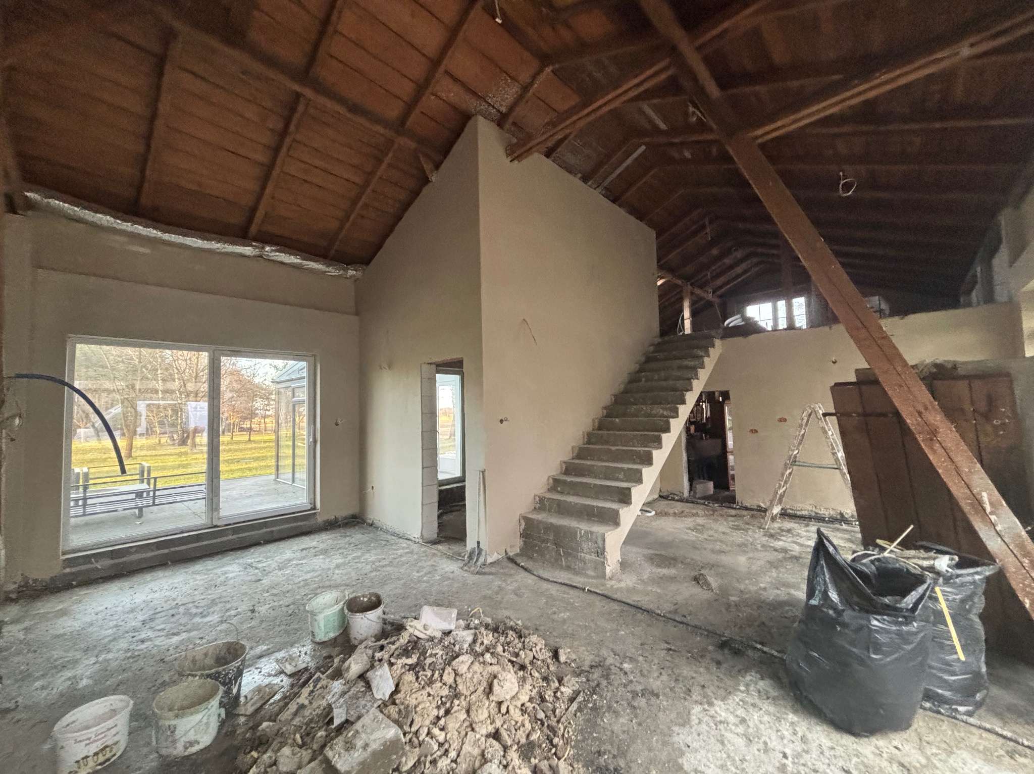 Surowe wnętrze domu w trakcie remontu, widoczne świeżo otynkowane ściany, betonowe schody, drewniany strop, okno z widokiem na zewnątrz, wiadra, gruz i worki na podłodze, drabina oparta o ścianę.